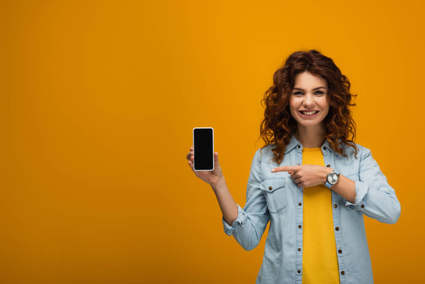 Veselá kudrnatá žena ukazující prstem na smartphone s prázdnou obrazovkou na oranžovém  - Fotografie, Obrázek