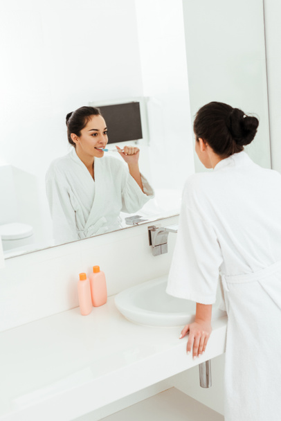 selective focus of happy young woman brushing teeth and looking at mirror in bathroom  - Foto, Bild