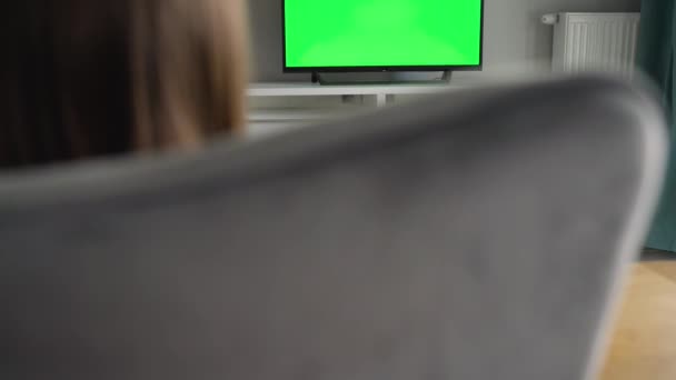Woman is sitting in a chair, watching TV with a green screen, switching channels with a remote control. Chroma key. Indoors - Footage, Video