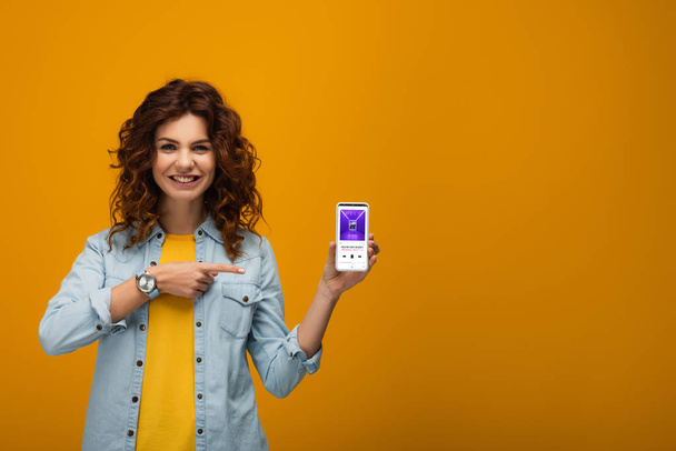 cheerful curly redhead woman pointing with finger at smartphone with itunes app on screen on orange  - Foto, Imagem