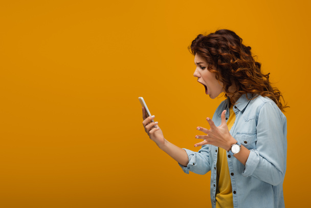 angry redhead woman gesturing while looking at smartphone and screaming on orange  - Fotó, kép