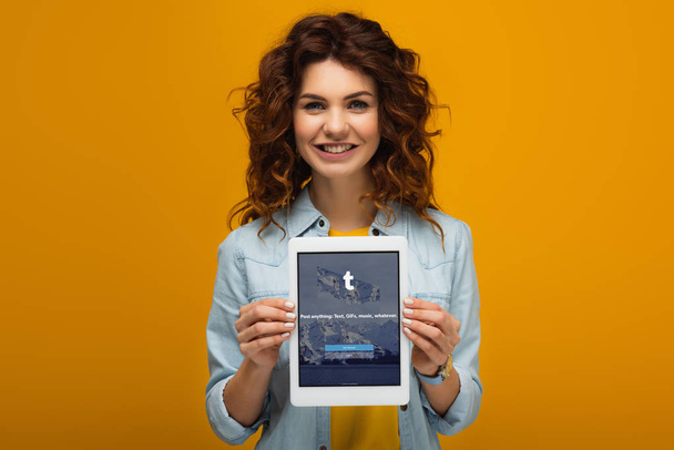fröhliche rothaarige Frau hält digitales Tablet mit Tumblr-App auf dem Bildschirm, während sie auf Orange steht  - Foto, Bild