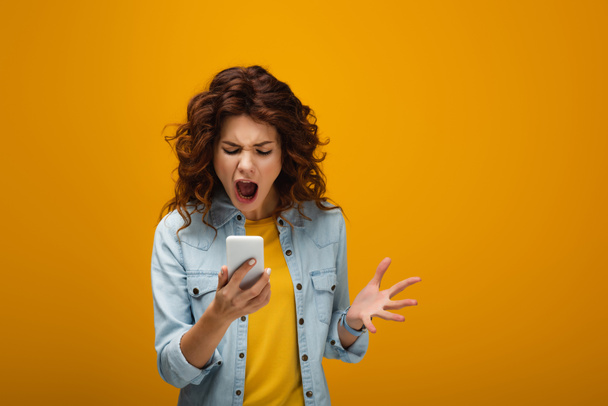 redhead woman gesturing while looking at smartphone and screaming on orange  - Fotó, kép