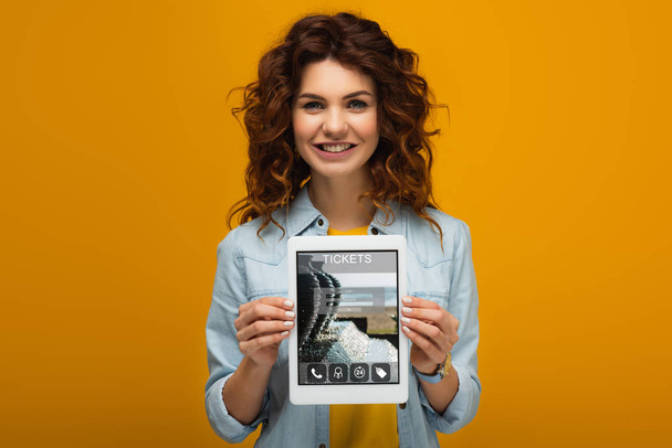 オレンジ色の上に立っている間、画面上のチケットアプリでデジタルタブレットを保持している幸せな赤毛の女性  - 写真・画像
