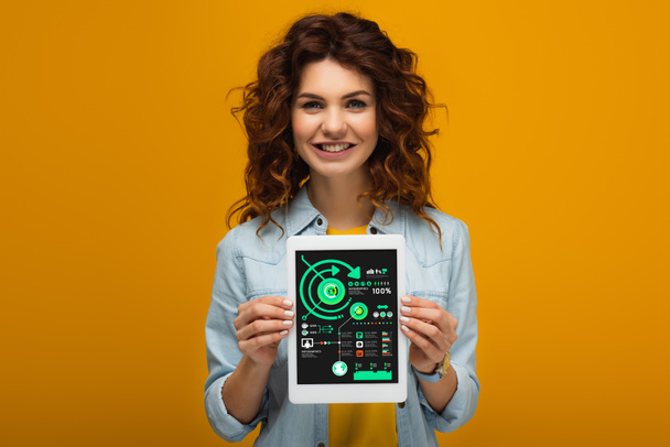 lächelnde rothaarige Frau hält ein digitales Tablet mit Diagrammen und Diagrammen auf dem Bildschirm, während sie auf Orange steht  - Foto, Bild
