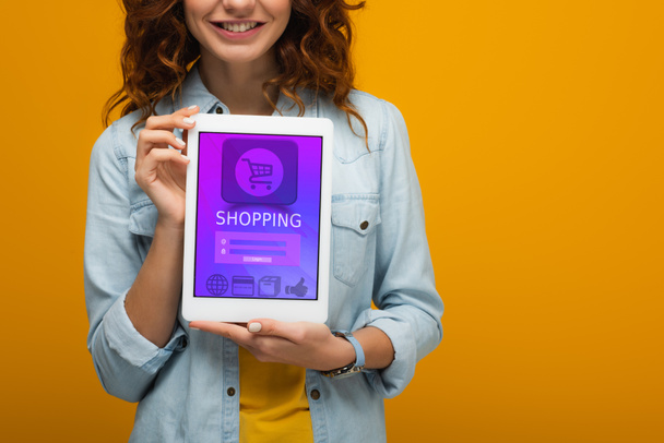 abgeschnittene Ansicht des fröhlichen lockigen Mädchens mit digitalem Tablet und E-Shopping-App auf dem Bildschirm isoliert auf orange  - Foto, Bild