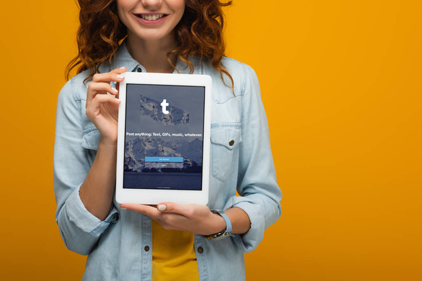 オレンジ色に隔離された画面上のタンブラーアプリでデジタルタブレットを保持する陽気なカーリーの女の子のトリミングビュー  - 写真・画像
