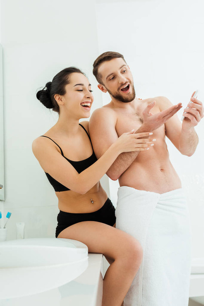 happy brunette woman in underwear looking at smartphone in hand of handsome boyfriend  - Photo, Image