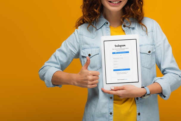 oříznutý pohled na veselou kudrnaté ženu držící digitální tablet s Instagram App na obrazovce a ukazující palec na oranžovou  - Fotografie, Obrázek