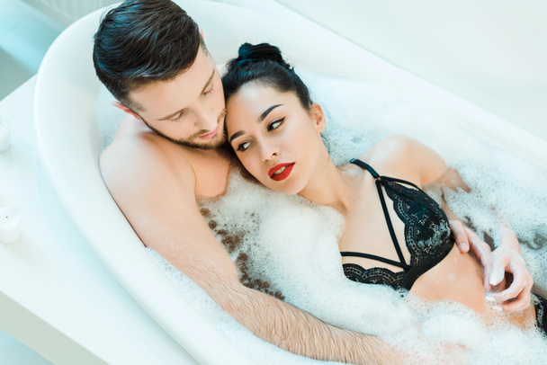 overhead view of handsome man lying in bathtub with attractive brunette woman in lace bra  - Photo, Image