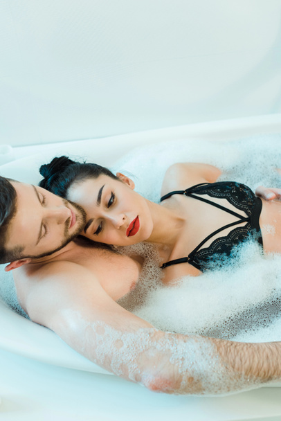 handsome shirtless man lying in bathtub with attractive brunette woman in lace bra  - Фото, изображение