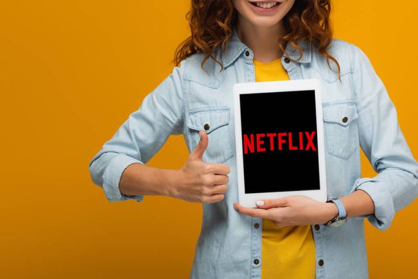 bijgesneden weergave van vrolijke krullend vrouw met digitale tablet met Netflix-app op het scherm en weergave van duim omhoog geïsoleerd op oranje  - Foto, afbeelding