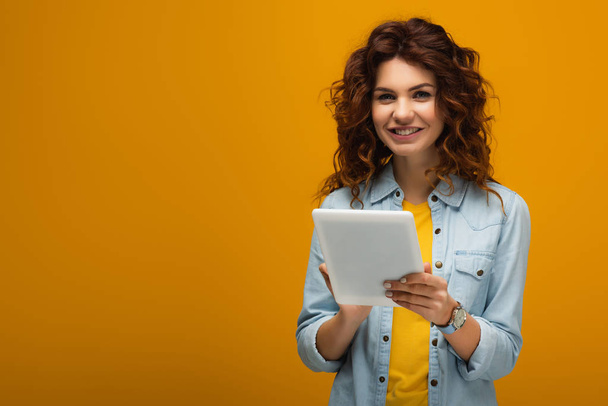 cheerful curly redhead girl looking at camera and holding digital tablet on orange  - Фото, зображення