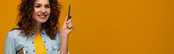 panoramic shot of smiling redhead young woman holding notebook and pen on orange  - Φωτογραφία, εικόνα