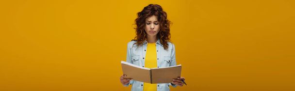 panoramic shot of upset redhead young woman looking at notebook and holding pen on orange  - Фото, изображение