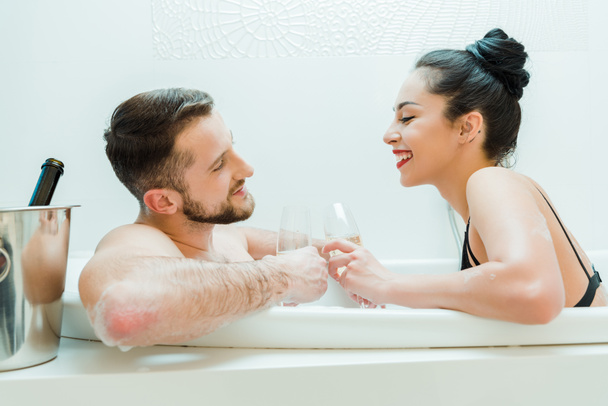 pohledný muž bez košile, který se dívá na šťastnou dívku držící ve vaně sklenici šampaňského  - Fotografie, Obrázek