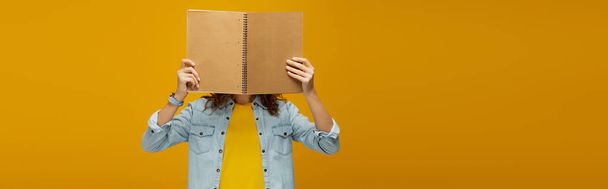 panoramic shot of young woman covering face with notebook on orange  - 写真・画像