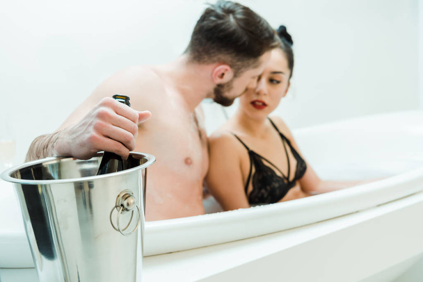 selective focus of man holding bottle of champagne near woman in bathtub  - Fotografie, Obrázek