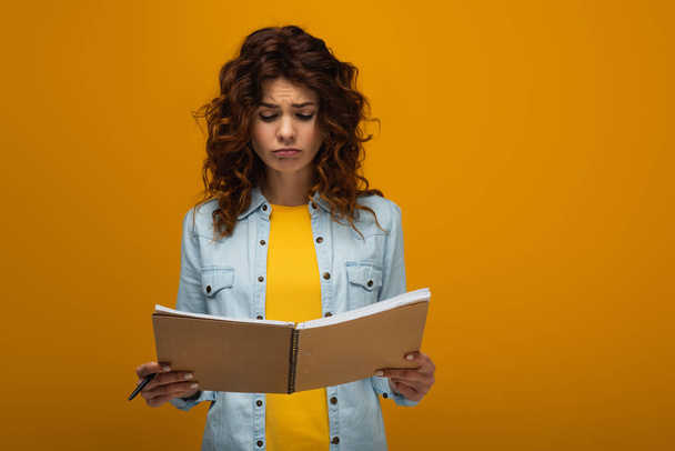 upset redhead young woman looking at notebook and holding pen on orange  - Foto, Bild