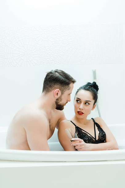 handsome shirtless man looking at brunette woman holding champagne glass in bathtub  - Фото, изображение
