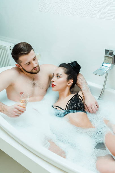 shirtless man hugging brunette woman and holding champagne glass in bathtub  - Фото, изображение