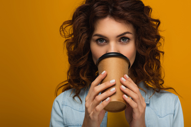 curly redhead woman covering face with paper cup while drinking isolated on orange  - Photo, Image
