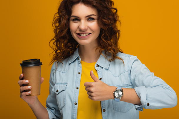 šťastná kudrnatá dívka, která drží kávu, aby šla a ukazovala palec na oranžovou  - Fotografie, Obrázek
