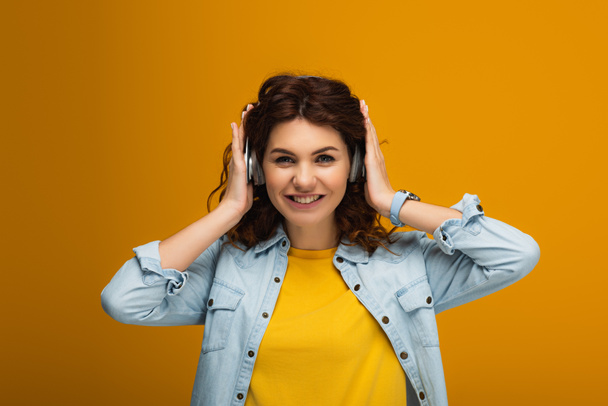 happy redhead girl listening music and touching headphones on orange  - Foto, Imagen