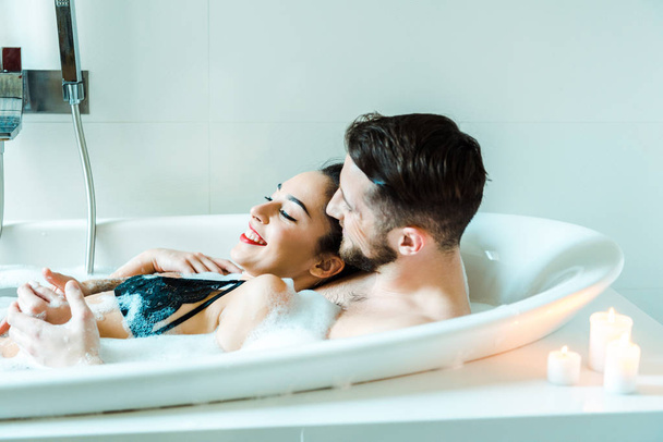 glückliche brünette junge Frau liegt mit bärtigem Mann in Badewanne  - Foto, Bild