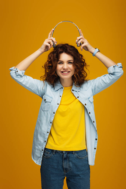 cheerful redhead girl smiling while holding headphones on orange  - Foto, Imagen