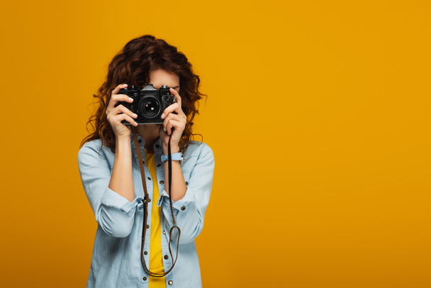 σγουρά κοκκινομάλλα φωτογράφος που καλύπτει το πρόσωπο με ψηφιακή φωτογραφική μηχανή απομονωμένη στο πορτοκαλί  - Φωτογραφία, εικόνα