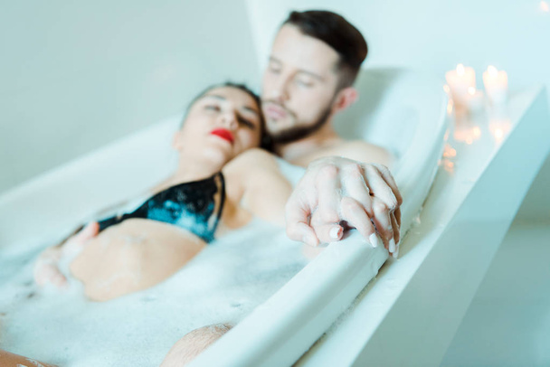 selective focus of handsome man and attractive brunette woman holding hands while lying in bathtub with bath foam  - Фото, изображение