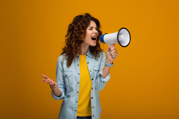 emotional curly redhead woman screaming in megaphone and gesturing on orange  - Foto, afbeelding