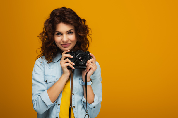 curly redhead photographer holding digital camera isolated on orange  - Zdjęcie, obraz
