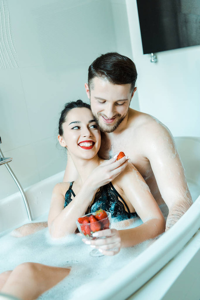 happy young woman holding tasty strawberry near cheerful boyfriend in bathtub  - Fotó, kép
