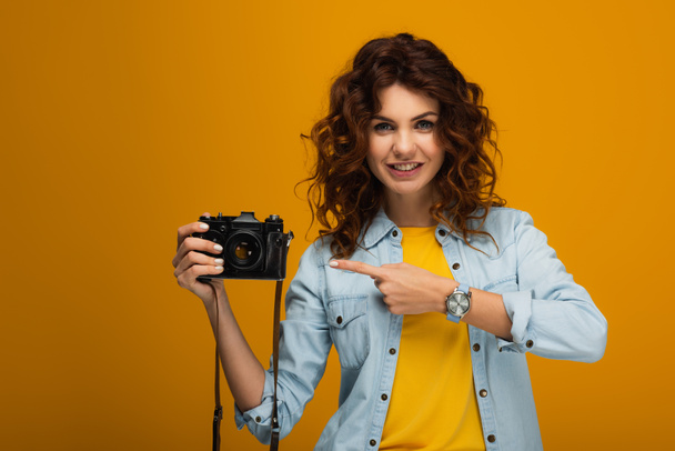 cheerful redhead photographer pointing with finger at digital camera on orange  - Φωτογραφία, εικόνα