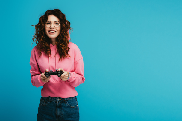 boldog göndör vöröshajú lány játszik videojáték, miközben tartja a joystick a kék - Fotó, kép