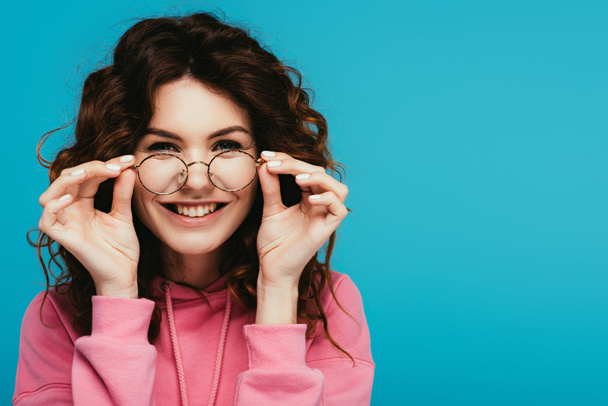 cheerful curly redhead girl touching glasses isolated on blue - Photo, Image