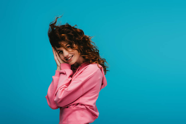 happy young woman with curly red hair looking at camera on blue  - Foto, Imagem