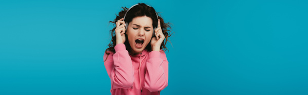 Panoramaaufnahme einer attraktiven, lockigen Frau, die singt, während sie Musik in Kopfhörern auf blau hört  - Foto, Bild