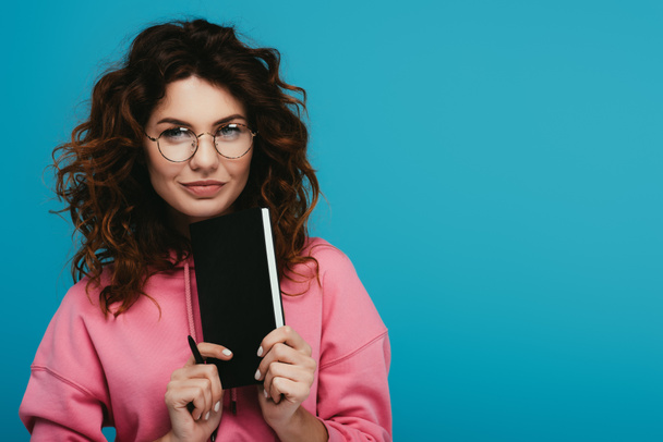 cheerful curly girl in glasses holding notebook and pen isolated on blue - Фото, зображення