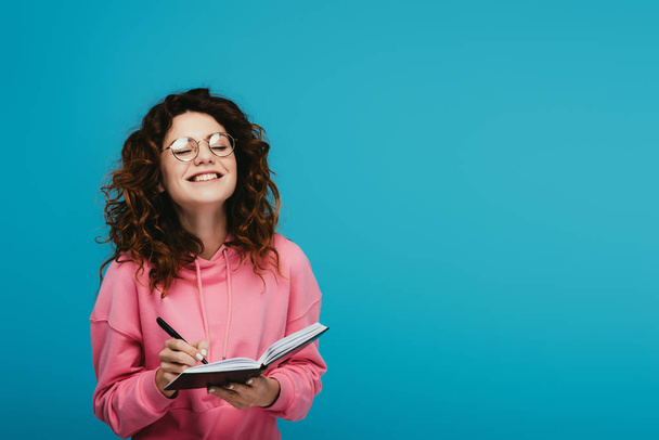 happy curly girl in glasses holding notebook and pen isolated on blue - Fotó, kép