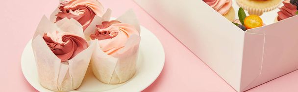 Panoramaaufnahme von leckeren Cupcakes auf weißem Teller und in Karton auf rosa Oberfläche - Foto, Bild