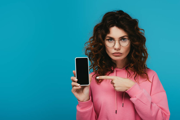 vonzó göndör vöröshajú lány mutat ujjal a smartphone üres képernyő elszigetelt kék  - Fotó, kép