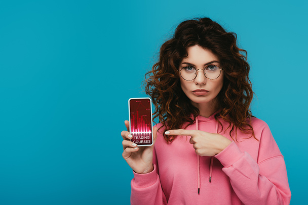 attraktive rothaarige Mädchen zeigt mit dem Finger auf das Smartphone mit Trading-Kurse auf dem Bildschirm isoliert auf blau  - Foto, Bild