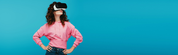 panoramic shot of curly girl wearing virtual reality headset while standing with hands on hips on blue  - Foto, imagen