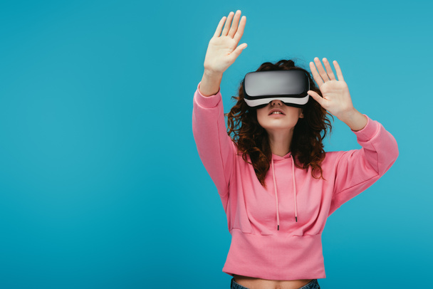 curly girl wearing virtual reality headset while gesturing on blue  - Foto, Imagen