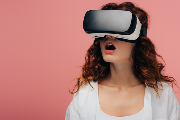 surprised curly redhead woman wearing virtual reality headset on pink  - Φωτογραφία, εικόνα