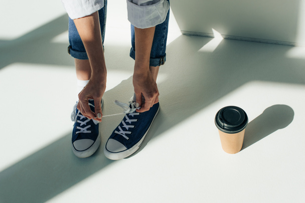 cropped view of woman tying shoelaces on sneakers while sitting near paper cup on white with sunshine  - Foto, immagini