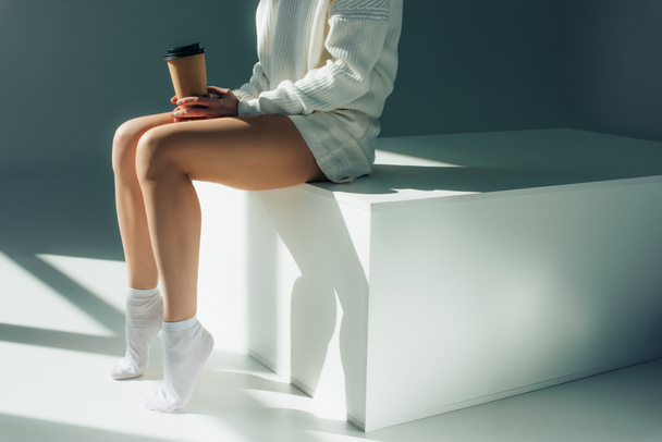 обрезанный вид молодой женщины, сидящей в носках и держащей бумажную чашку на сером
 - Фото, изображение